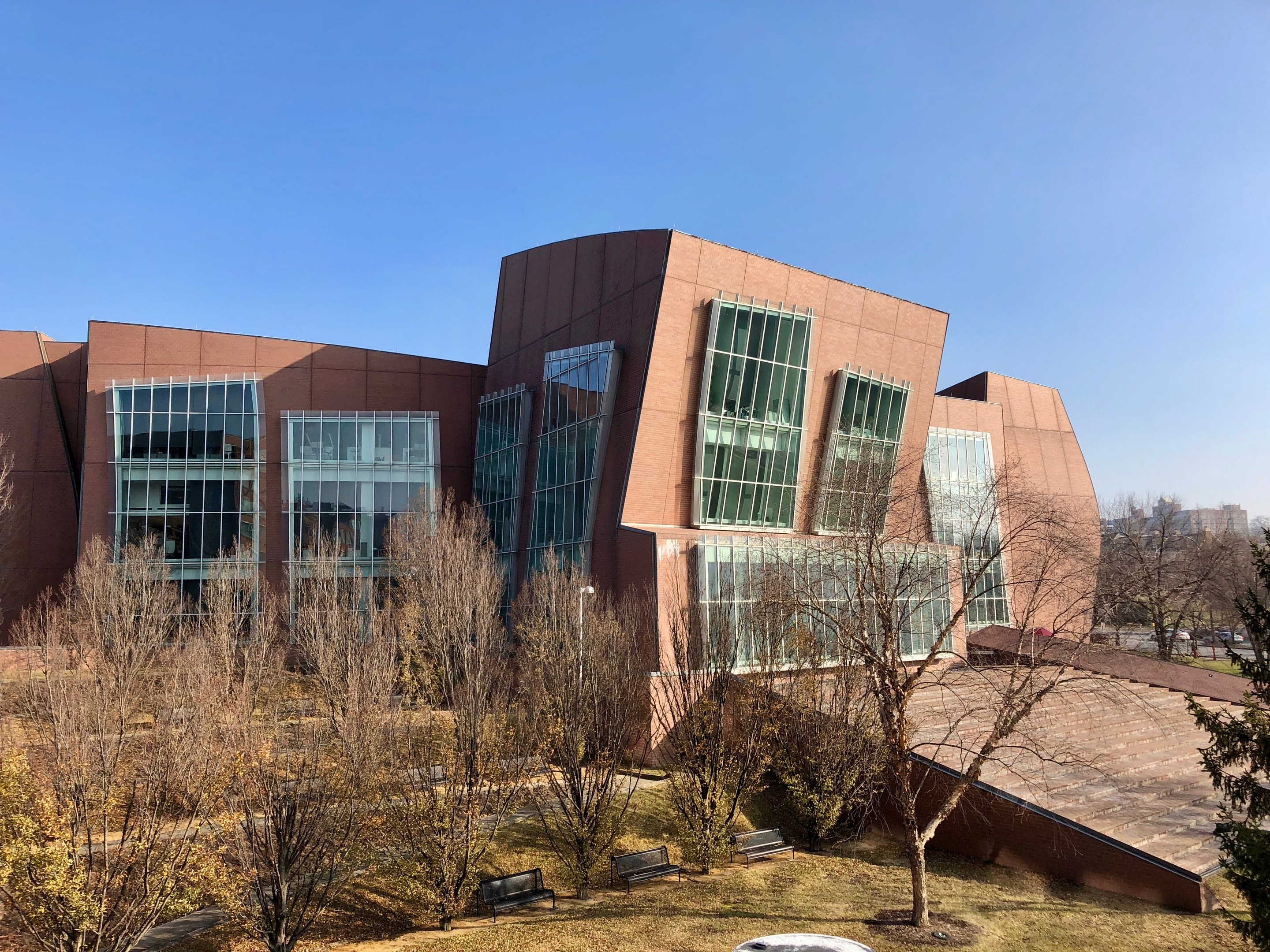 Großes, mehrstöckiges Gebäude mit zahlreichen Fenstern, umgeben von Bäumen, Bänken, Gras und einem Lichtmast, mit anderen Gebäuden und einem klaren blauen Himmel im Hintergrund.