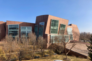 Großes, mehrstöckiges Gebäude mit zahlreichen Fenstern, umgeben von Bäumen, Bänken, Gras und einem Lichtmast, mit anderen Gebäuden und einem klaren blauen Himmel im Hintergrund.