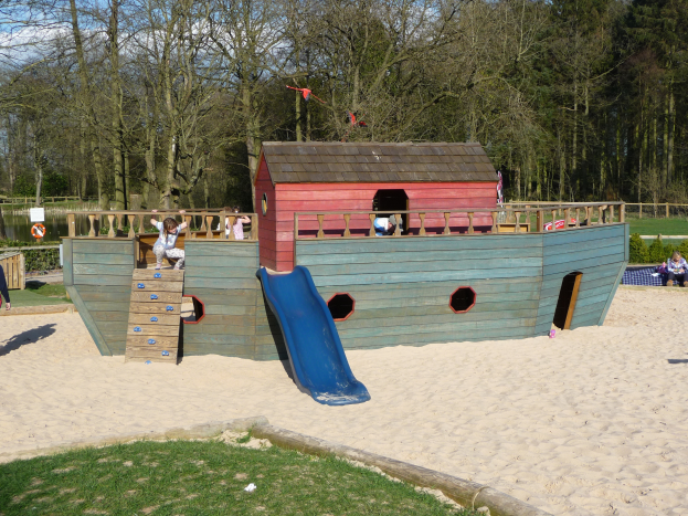 Ein Piratenschiff-Spielhaus mit einer Rutsche in der Mitte, umgeben von Sand und Gras, mit spielenden Menschen und Bäumen sowie einem klaren blauen Himmel im Hintergrund.
