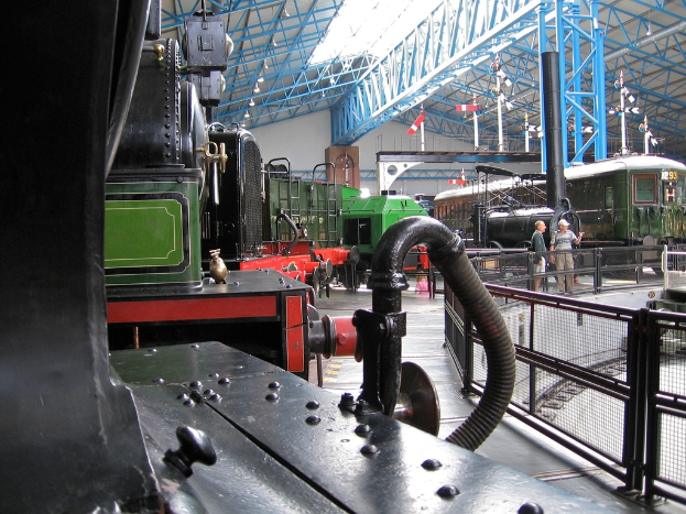 Mehrere Zugmaschinen sind in einem Abstellgleis mit zahlreichen Eisenstangen oben geparkt. Rechts verläuft ein Zaun entlang einer Bahnstrecke mit einem Zug. Zwei Personen stehen in der Mitte der Szene nahe dem Zaun.