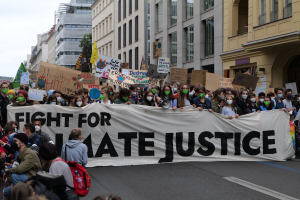 Eine große Gruppe von Menschen, die eine Straße entlangmarschieren und ein Banner mit der Aufschrift "Kämpfen für Hassjustiz" tragen, einige tragen Masken und halten Schilder, und im Hintergrund Gebäude, Bäume und einen klaren blauen Himmel.