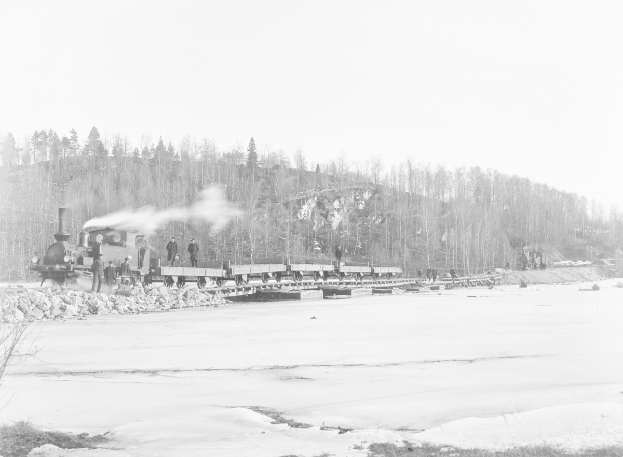 Ein Schwarz-Weiß-Foto eines Zuges, der über einen zugefrorenen Fluss fährt, mit Menschen auf einer Brücke, Bäumen, einem Hügel und dem Himmel im Hintergrund.