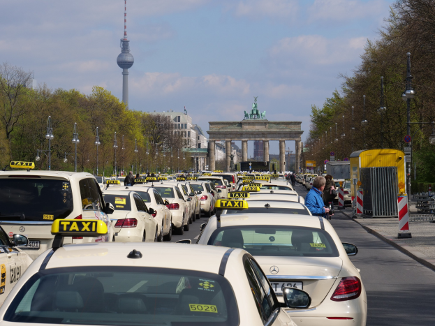 Eine belebte Straße in Berlin mit zahlreichen geparkten Taxis, Fußgängern auf dem Gehweg, Laternen, Bäumen, Gebäuden, einem fernen Bogen mit Statuen und einem Turm sowie einem bewölkten Himmel.