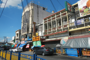 Eine belebte Stadtstraße mit Autos, hohen Gebäuden, Schildern, Regenschirmen, einem Metallzaun, Strommasten mit Kabeln und einem bewölkten Himmel.