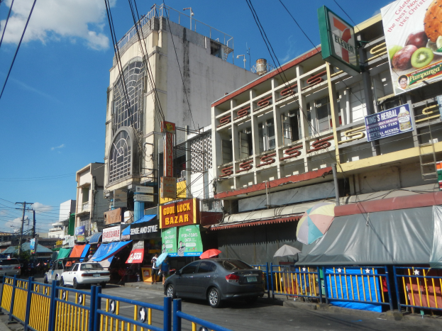 Eine belebte Stadtstraße mit Autos, hohen Gebäuden, Schildern, Regenschirmen, einem Metallzaun, Strommasten mit Kabeln und einem bewölkten Himmel.