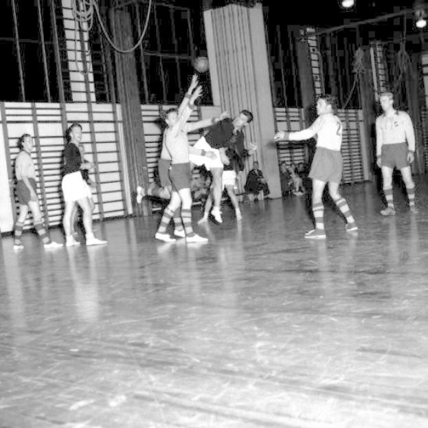Eine Gruppe von Männern, die Handball auf einem Holzfußboden spielen, mit einer Wand und Scheinwerfern im Hintergrund, in Schwarz-Weiß.