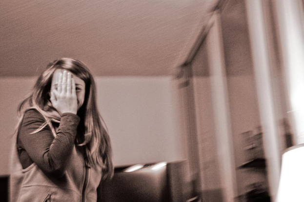 Eine Frau steht auf der linken Seite des Bildes, mit einem Glasfenster auf der rechten Seite, einer Wand im Hintergrund, einer Decke oben, einer Lampe unten und einer Couch in der Szene.