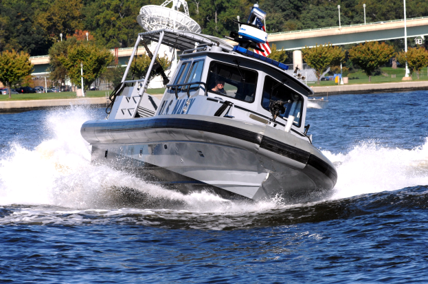 Ein Polizeiboot fährt über einen Gewässser mit einer Person darin, mit einer Brücke, Bäumen, Pfählen, Fahrzeugen, Gras und einem klaren blauen Himmel im Hintergrund.