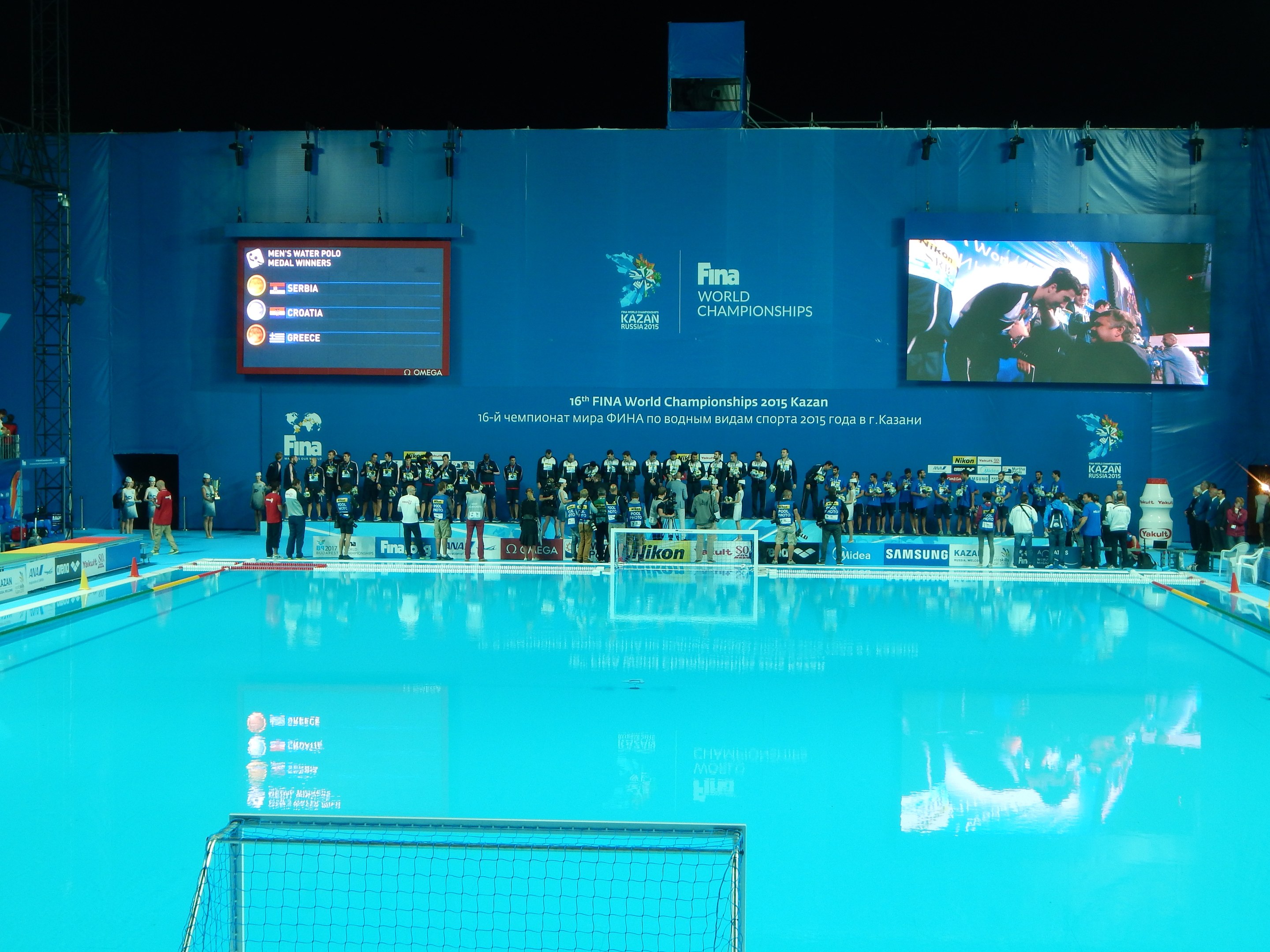 Gruppe von Menschen um ein Hallenbad mit Netz am Boden herumstehend, mit einer Wand mit "FINA World Championships 2015 Kazan"-Text und -Logos, Trägern und Lampen im Hintergrund.