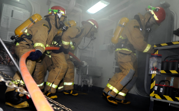 Feuerwehrleute in Schutzausrüstung, darunter Helme und Sauerstoffflaschen, arbeiten an einem Feuerwehrauto, mit einem Gestell voller verschiedener Gegenstände auf der rechten Seite und Rohren, Lichtern und anderen Gegenständen im Hintergrund.