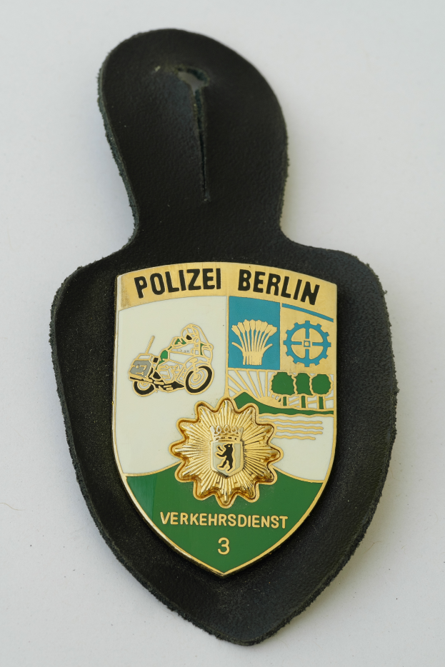 Ein Polizeistern mit der Aufschrift "Polizei Berlin" darauf, das ein blaues Schild mit einem weißen Stern in der Mitte zeigt, umgeben von einem weißen Rand, auf einer Oberfläche platziert.