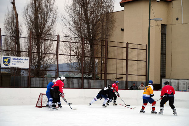 Menschen, die Eis hockey auf einem Eisplatz spielen, mit Gebäuden, Bäumen, einer Straßenlaterne, einem Namensschild und Zäunen im Hintergrund unter einem Himmel.