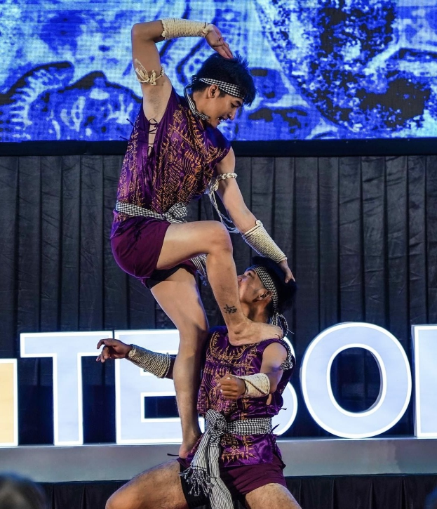 Zwei Performer in lila Kleidern führen einen akrobatischen Hebeflug auf der Bühne vor einem Publikum aus, mit schwarzem Vorhang und Bildschirmhintergrund.