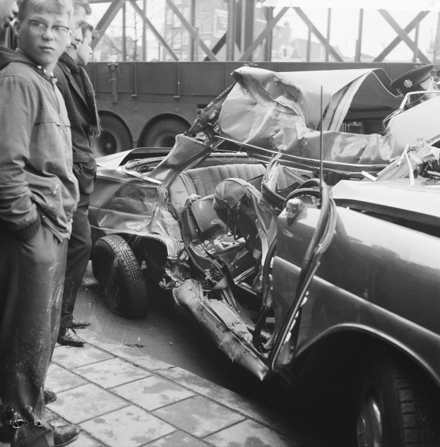 Schwarz-weiß-Bild eines verunglückten Autos am Straßenrand mit Menschen in der Nähe, einem Lkw und einer Brücke im Hintergrund.