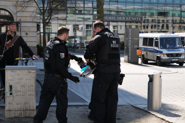 Drei Polizeibeamte stehen zusammen auf einer Stadtstraße, zwei in schwarzen Uniformen und einer mit einem Fahrrad, mit Fahrzeugen, Bäumen und Gebäuden im Hintergrund.