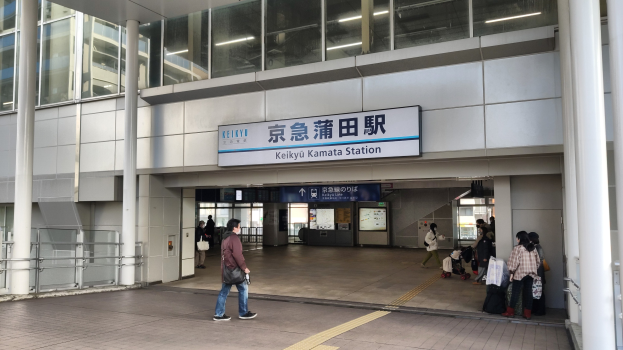 Menschen mit Taschen durch eine Bahnhofshalle gehen, ein zentraler Mann in Fußbekleidung, ein Textschild, Pfeiler, Geländer, Glaswände und Deckenleuchten.