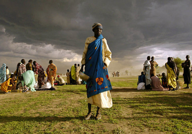 Eine Frau in einem blauen Kleid steht mit einem silbernen Gefäß in der Hand und wird von einer Gruppe Frauen umgeben, die hinter ihr sitzen und stehen, unter einem bewölkten Himmel mit Bäumen im Hintergrund.