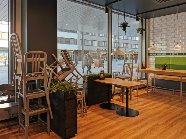 Ein Restaurantinnenraum mit hölzernen Tischen und Stühlen, großen Fenstern, die eine städtische Straßenszene mit Menschen, Gebäuden und Himmel zeigen, umgeben von Pflanzen und Lichtern.