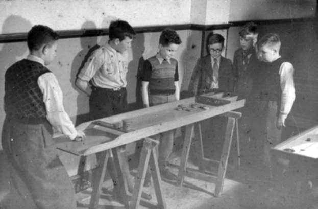 Eine Gruppe von Jungs, die um einen Tisch stehen und Tischtennis spielen, in einem Raum mit einer Wand im Hintergrund, in Schwarz-Weiß dargestellt.