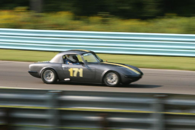 Ein Auto fährt auf einer Straße mit einer Person mit Helm im Inneren, Absperrungen auf beiden Seiten und Bäumen im Hintergrund.