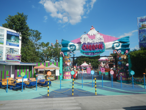 Eine Karussellfahrt am Eingang eines Parks mit einer Straße, einem eingezäunten Bereich, Buden, Fahnen, Bäumen und einem bewölkten Himmel im Hintergrund.