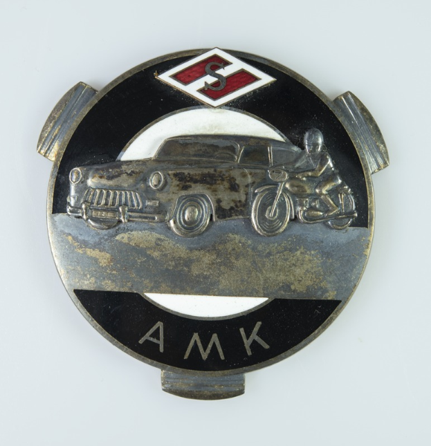 Ein schwarzes und silbernes Abzeichen mit einem zentralen Auto, einem Motorrad rechts daneben und dem Text "AMK" darüber, alles vor einem weißen Hintergrund.