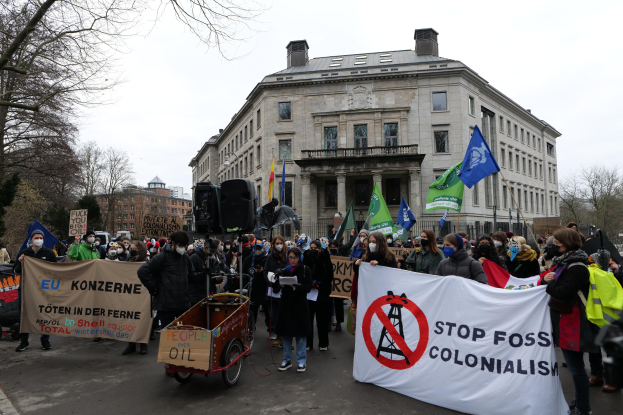 Große Gruppe von Menschen marschiert bei einer Demonstration gegen fossile Brennstoffe, trägt Schilder und Fahnen, mit einem Fahrzeug im Vordergrund und Gebäuden, Bäumen und einem klaren Himmel im Hintergrund.
