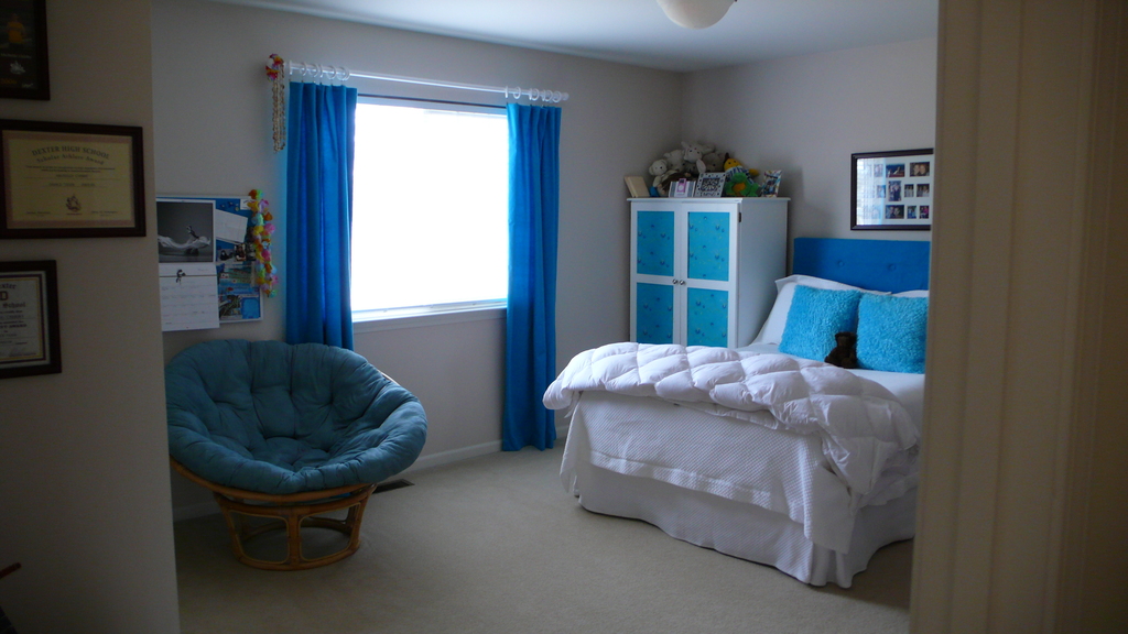 Ein Raum mit einem Bett, Stuhl, Fenster mit Vorhängen, Schrank, Foto Rahmen und einem gefliesten Boden.