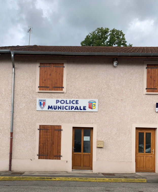 Ein Gebäude der Polizei municipale mit einem Schild, Fenstern, Türen, einer Namensplakette, einem Rohr, einem Baum und einem bewölkten Himmel im Hintergrund.