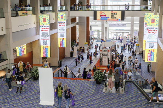 Eine große Gruppe von Menschen, die durch ein großes Gebäude mit Bannern, Topfpflanzen, Rolltreppen, Geländern, Säulen, Texttafeln, Glastüren und anderen Gegenständen geht, einige tragen Taschen bei der Nationalen Buchmesse.