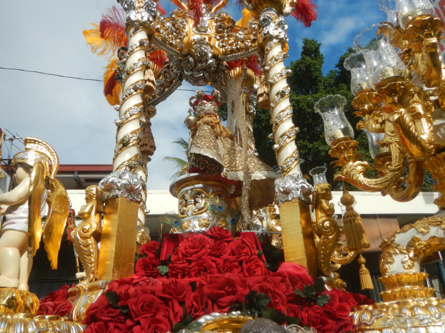 Ein Rosenmontagszug-Schmuckwagen mit roten Rosen und goldenen Verzierungen, darunter Statuen und Blumen, mit einem Gebäude, Bäumen und einem bewölkten Himmel im Hintergrund.