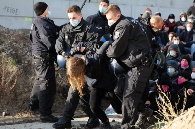 Gruppe maskierter Personen in Mützen, einige mit Waffen, umringt eine Frau, die von der Polizei eskortiert wird, mit Pflanzen, Steinen und einer Wand mit Protesttext im Hintergrund.