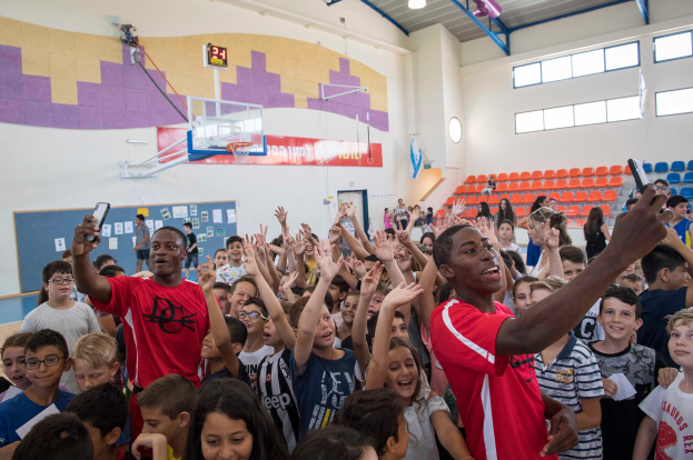Eine Gruppe von Kindern steht vor einem Basketballfeld, einige halten Mobiltelefone, mit einer Tafel, Uhr, Torpfosten, Basketballkorb, Deckenleuchten, Stühlen und Fenstern im Hintergrund.