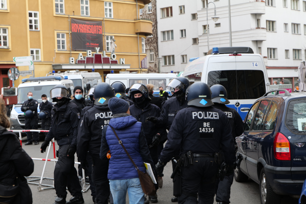 Eine Gruppe von Polizisten steht vor einer Menge von Menschen, die Helme und Jacken tragen, mit Fahrzeugen auf der Straße und einer Absperrung im Vordergrund, während im Hintergrund Gebäude, Laternenpfähle, Texttafeln, ein Banner und eine Statue zu sehen sind.