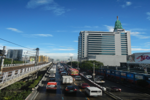 Eine belebte Stadtstraße mit starkem Verkehr, hohen Gebäuden, einer Brücke links, Strommasten mit Drähten, Bäumen und einem bewölkten Himmel im Hintergrund.
