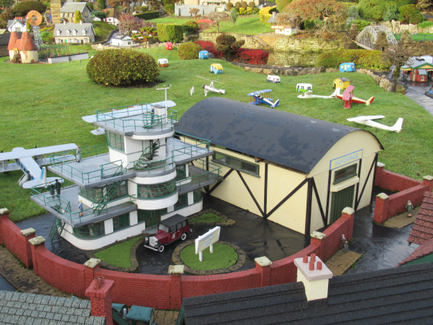 Ein detailliertes Modell einer Kleinstadt mit verschiedenen Gebäuden, Bäumen, Pflanzen, Fahrzeugen, Flugzeugen, Menschen und anderen Objekten, mit Häusern am Boden, die einen belebten Eindruck einer Stadt erwecken.