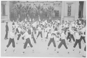 Eine Gruppe von Männern in weißen Hemden und schwarzen Hosen trainiert Boxen vor einem Gebäude mit Fenstern und Bäumen im Hintergrund, dargestellt in Schwarz-Weiß.