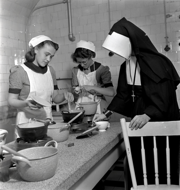 Drei Frauen in Schürzen und Häubchen stehen in einer schwarz-weißen Küche und bereiten Essen mit Utensilien und Schüsseln auf der Arbeitsfläche vor, rechts daneben ein Stuhl und im Hintergrund Rohre.