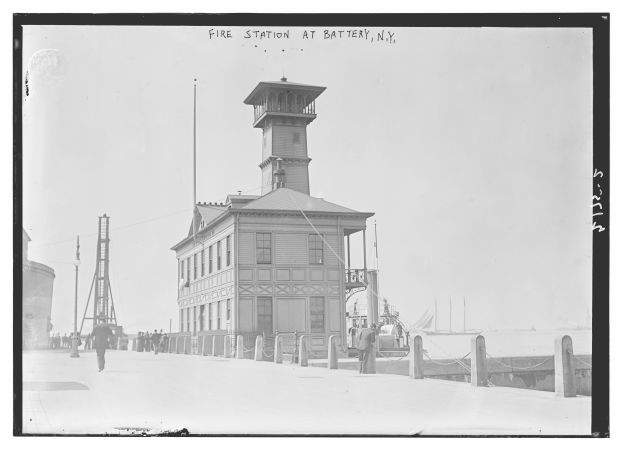 Ein Schwarz-Weiß-Foto einer Feuerwache in Battery, New York, mit einem Gebäude, Menschen am Boden, Pfählen, einem Zaun, Booten auf dem Wasser und einer bewölkten Himmel.