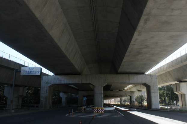 Autobahn unter einer Brücke mit Absperrkegeln, Pollern und Texttafeln, flankiert von Geländern, Bäumen und einem klaren blauen Himmel im Hintergrund.