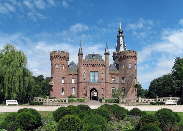 Eine Burg mit mehreren Zinnen steht in einem Park mit grünem Gras, Pflanzen, Bäumen, Bänken davor und sichtbaren Fenstern und Türen unter einem teilweise bewölkten Himmel.