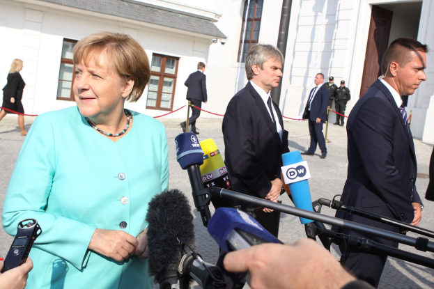 Bundeskanzlerin Angela Merkel beim Sprechen mit Reportern vor dem Kanzleramt in Berlin, umgeben von einer Gruppe von Menschen, einige halten Mikrofone, mit Gebäuden im Hintergrund.