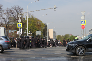 Eine Gruppe von Polizisten in schwarzen Uniformen und Helmen steht in der Mitte einer Straße mit Autos, Laternenmasten, Schildern, Bäumen, Gebäuden und einem klaren blauen Himmel im Hintergrund.