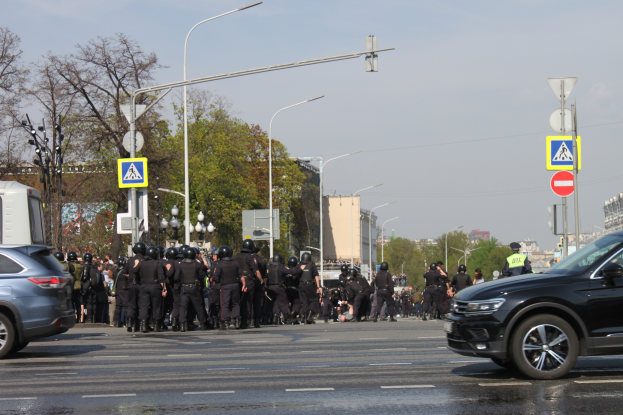 Eine Gruppe von Polizisten in schwarzen Uniformen und Helmen steht in der Mitte einer Straße mit Autos, Laternenmasten, Schildern, Bäumen, Gebäuden und einem klaren blauen Himmel im Hintergrund.