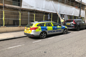 Ein Polizeiwagen steht an der Seite einer Straße neben einem Gebäude, zwei Helme tragende Personen stehen daneben, ein Geländer und ein Pfahl sind im Vordergrund zu sehen, und ein weißes Tuch hängt über das Gebäude im Hintergrund, was auf eine polizeiliche Untersuchung nach einem Messerangriff in London hindeutet.