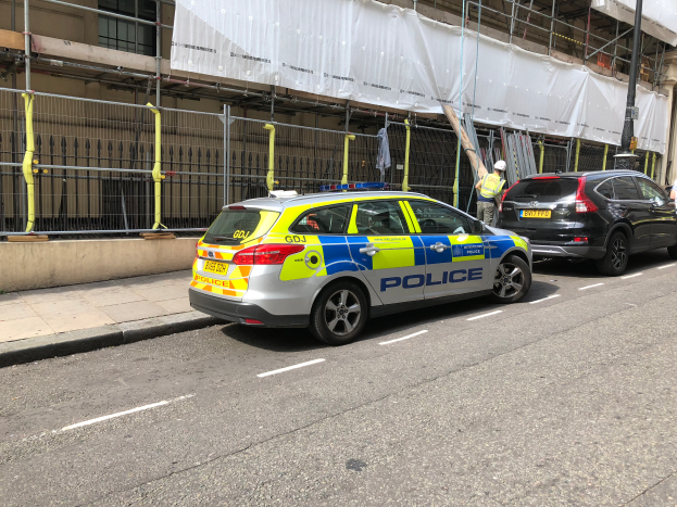 Ein Polizeiwagen steht an der Seite einer Straße neben einem Gebäude, zwei Helme tragende Personen stehen daneben, ein Geländer und ein Pfahl sind im Vordergrund zu sehen, und ein weißes Tuch hängt über das Gebäude im Hintergrund, was auf eine polizeiliche Untersuchung nach einem Messerangriff in London hindeutet.