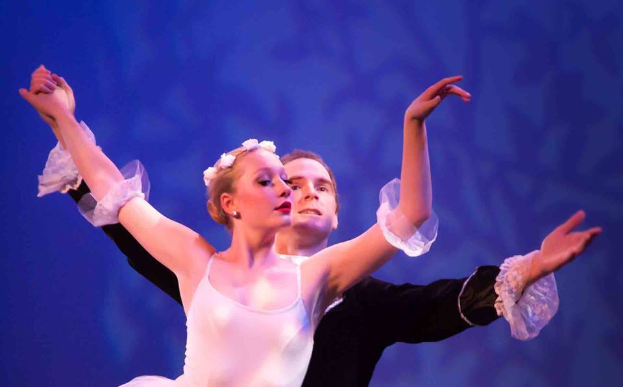 Ein Mann in einem schwarzen Kleid und eine Frau in einem weißen Kleid tanzen anmutig auf einer Bühne mit blauem Hintergrund und schauen sich verliebt und bewundernd in die Augen während einer Ballettvorstellung.