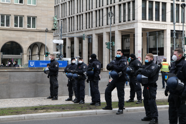 Eine Gruppe von Polizisten in schwarzen Uniformen und Masken steht vor einem Gebäude mit Glasfenstern und Säulen in Berlin, Deutschland, einige halten Helme, mit Laternen, Verkehrszeichen, Tafeln mit Text, einer Statue, anderen Menschen und einem grasbewachsenen Boden im Hintergrund.