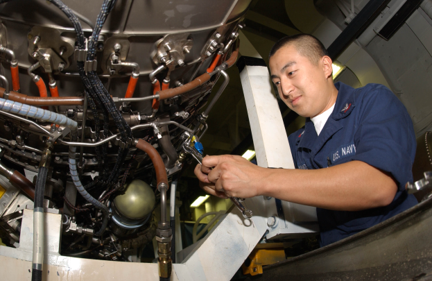 Ein Techniker in blauer Uniform arbeitet an einem Flugzeugtriebwerk in einer Fabrik und hält ein Werkzeug in der Hand.