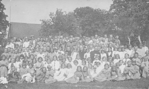 Schwarz-weiß-Foto einer Gruppe von Kindern, die vor einem Haus mit Bäumen und einem klaren Himmel posen.